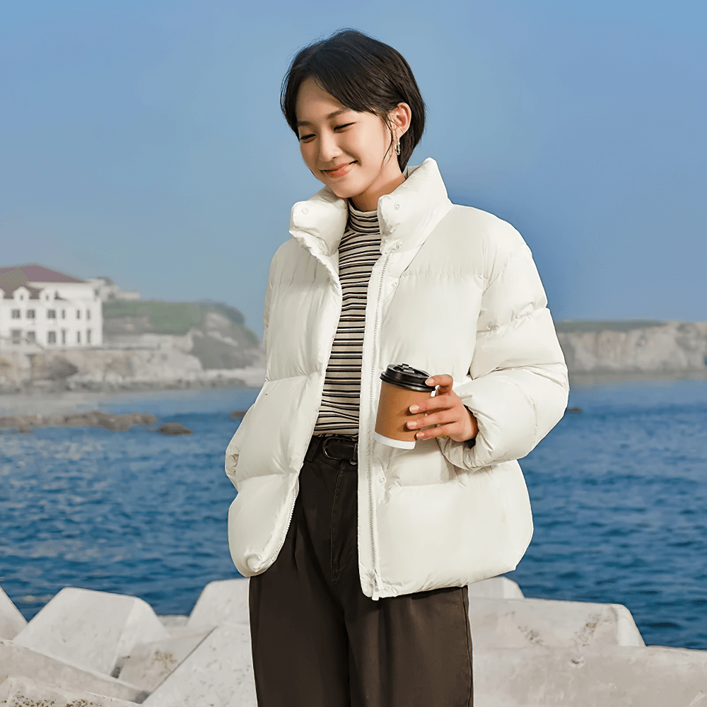 Woman wearing white goose down jacket with stand-up collar by the sea, holding a cup. Casual winter coat fashion for women.