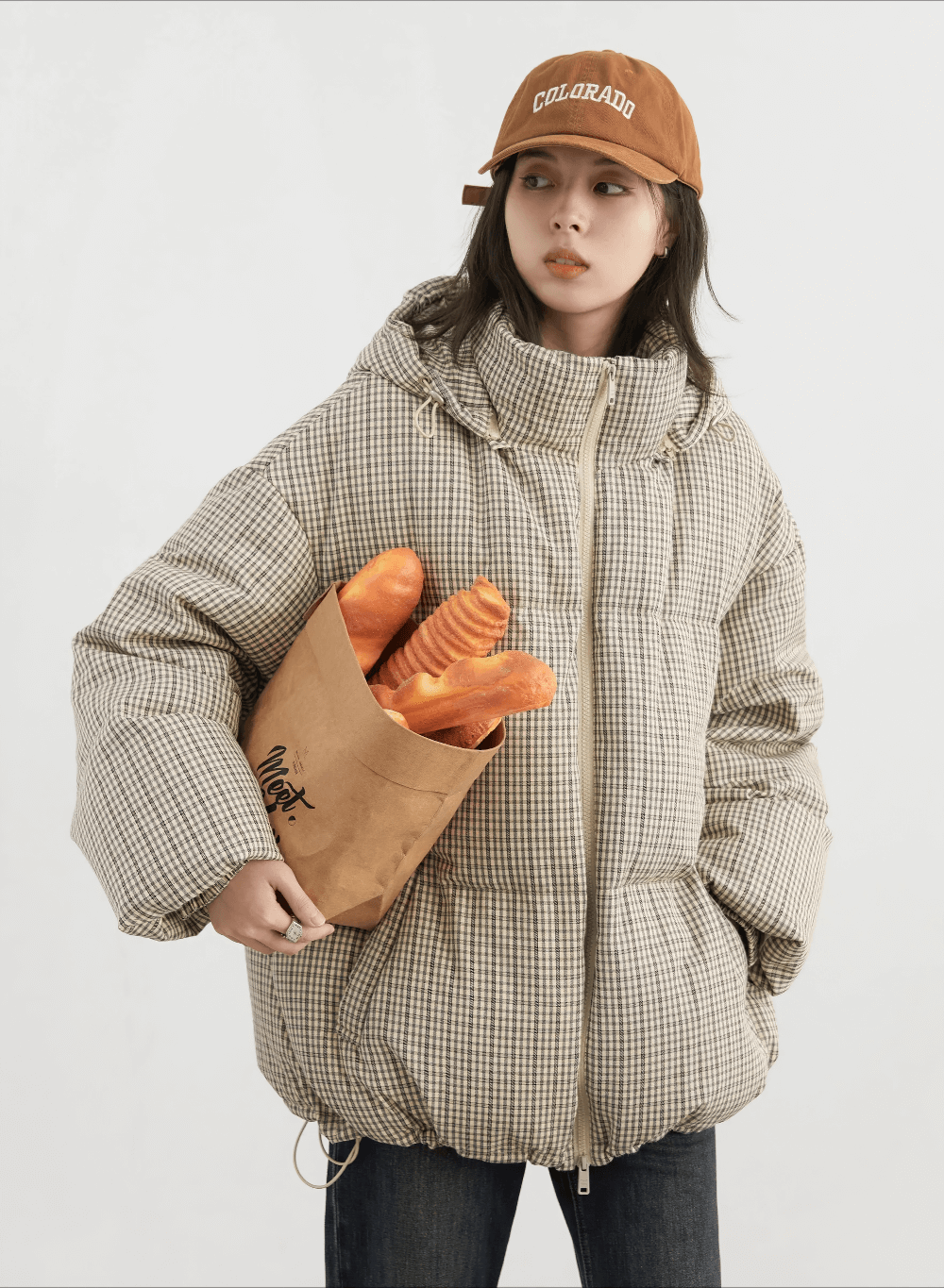 Woman in plaid hooded down jacket with hood, holding a bag of bread, wearing a cap, showcasing winter fashion and duck down insulation.