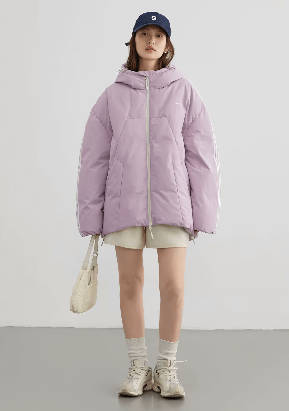 Woman in lavender hooded down jacket with white stripes and casual streetwear look, holding a bag, wearing a cap and sneakers.