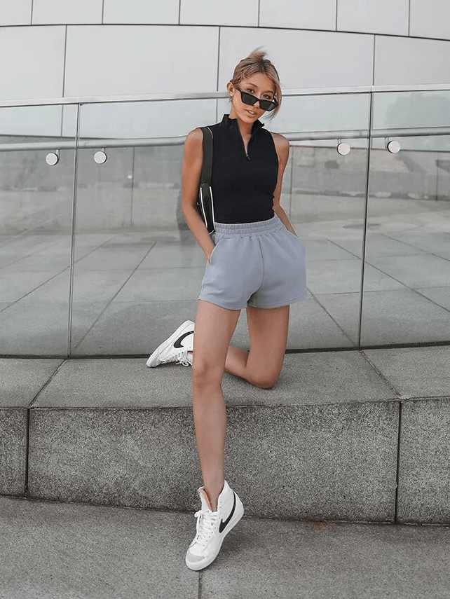 Woman wearing stylish black top, gray shorts, and white sneakers, leaning casually against a modern glass railing.