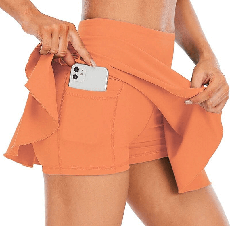 Woman wearing high waist tennis skirt-shorts with a pocket, holding an orange themed smartphone, showcasing breathable sportswear style.