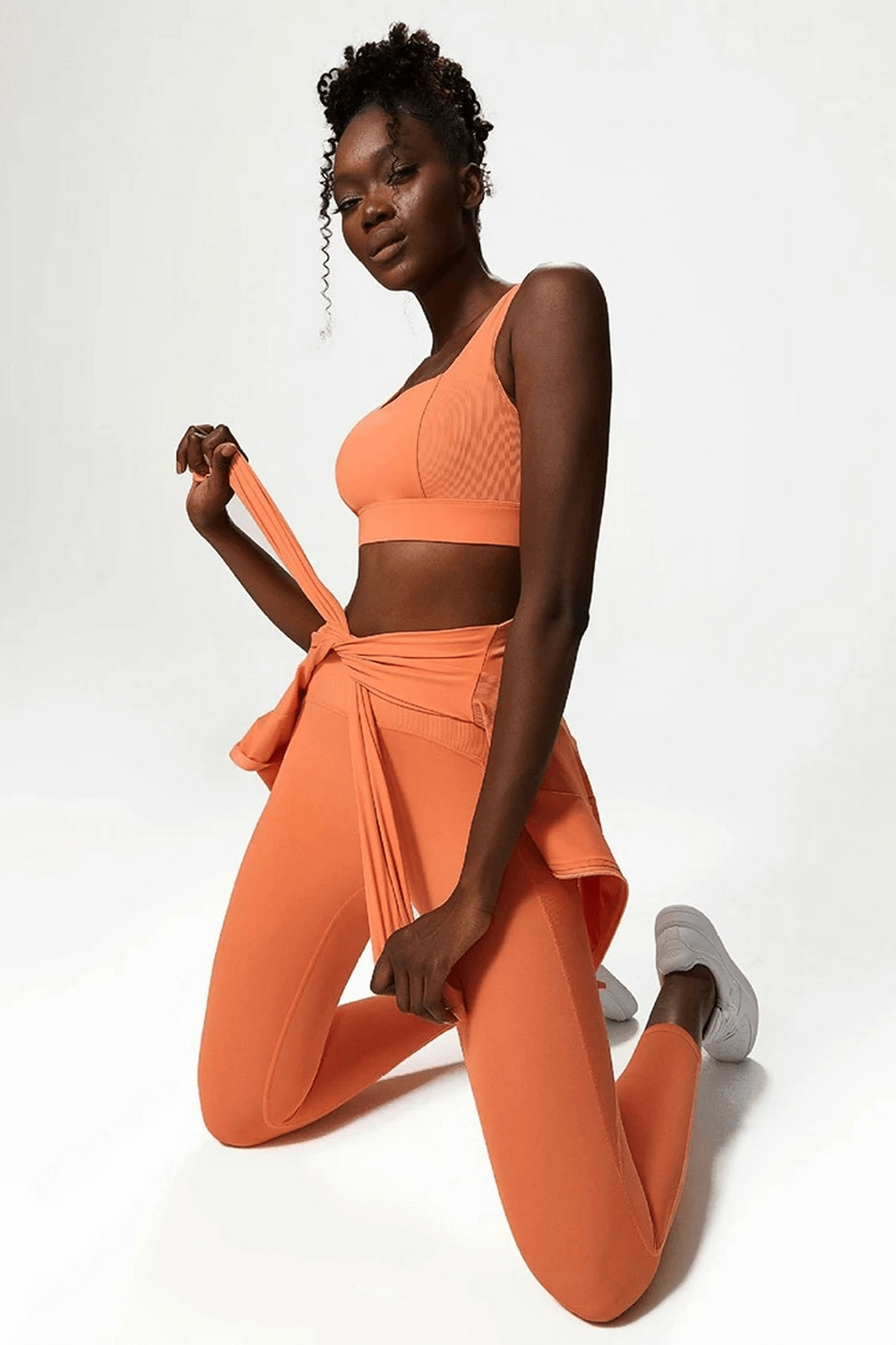 Woman in orange yoga leggings and sports bra kneeling, showcasing stylish nylon-spandex activewear for gym and yoga. Model exudes confidence.