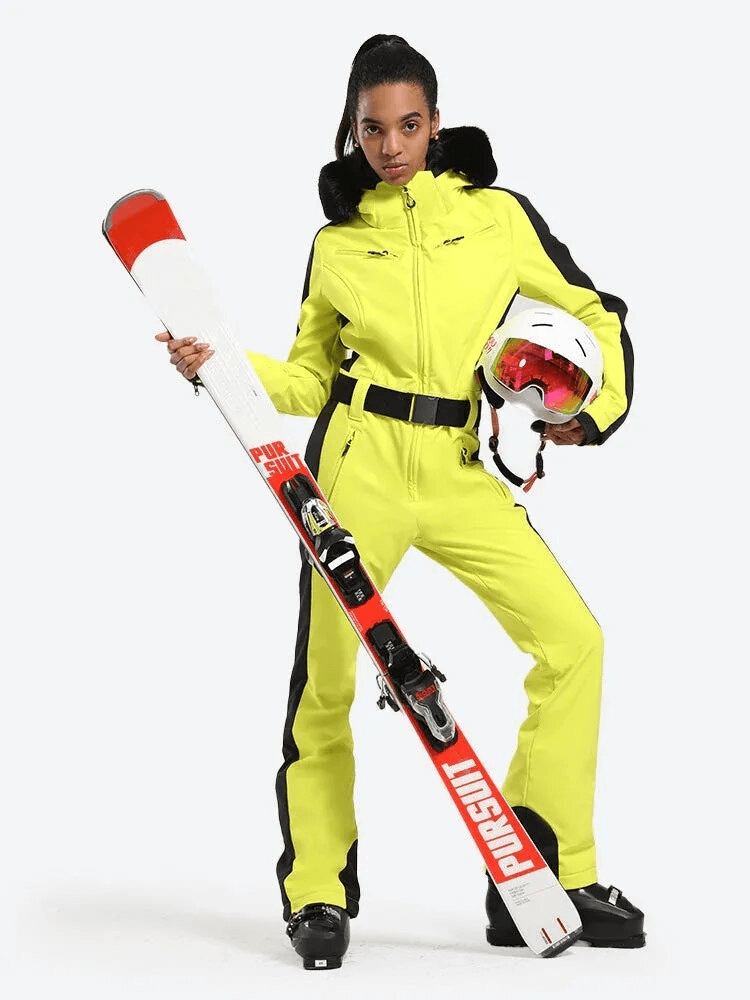 Woman in yellow one-piece ski suit with hood holding skis and helmet.