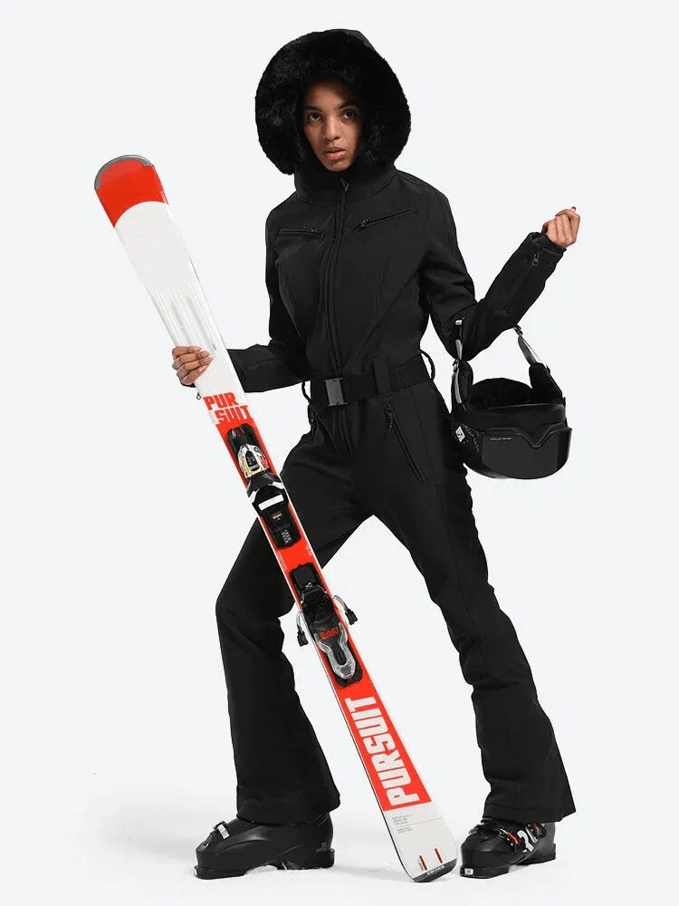 Stylish woman in black one-piece ski suit with hood, holding skis, ready for mountain adventure.