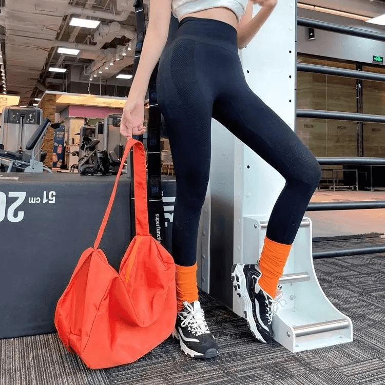 Woman at the gym with a large-capacity waterproof orange tote bag and sporty leggings, showcasing trendy fitness style.