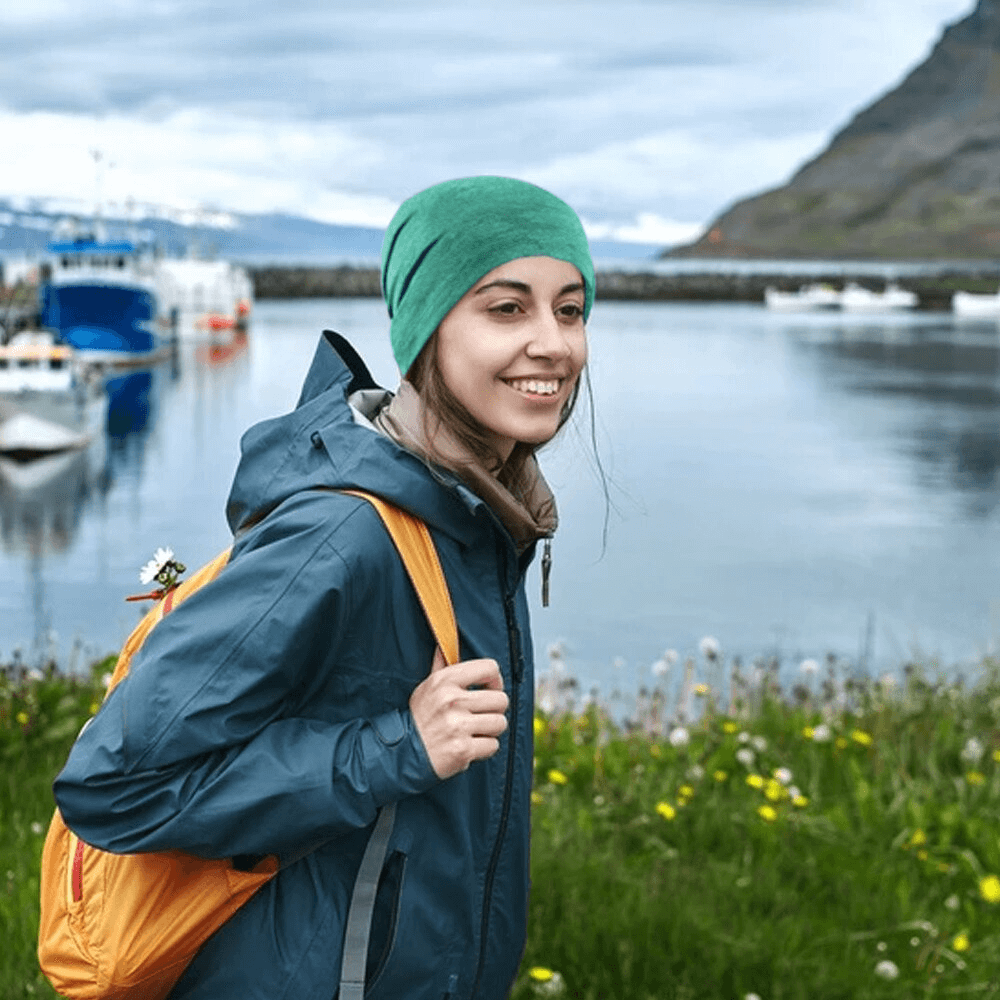 Woman wearing a green breathable running hat, standing by a lakeside, dressed for hiking with a blue jacket and yellow backpack.