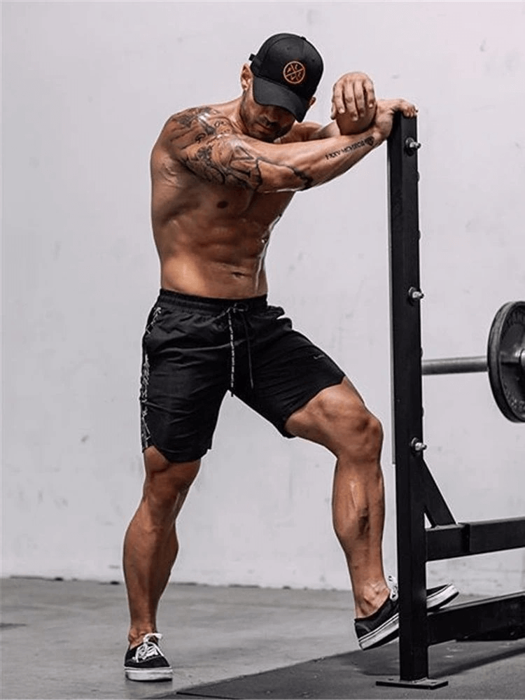 Athlete wearing drawstring black running shorts, exercising at the gym by a weight rack, showcasing bodybuilding gym attire.
