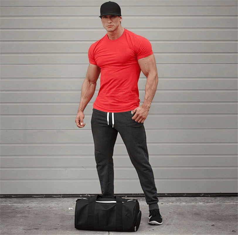 Man wearing red quick-dry sports T-shirt and black cap, standing with a black duffel bag in front of a gray background.