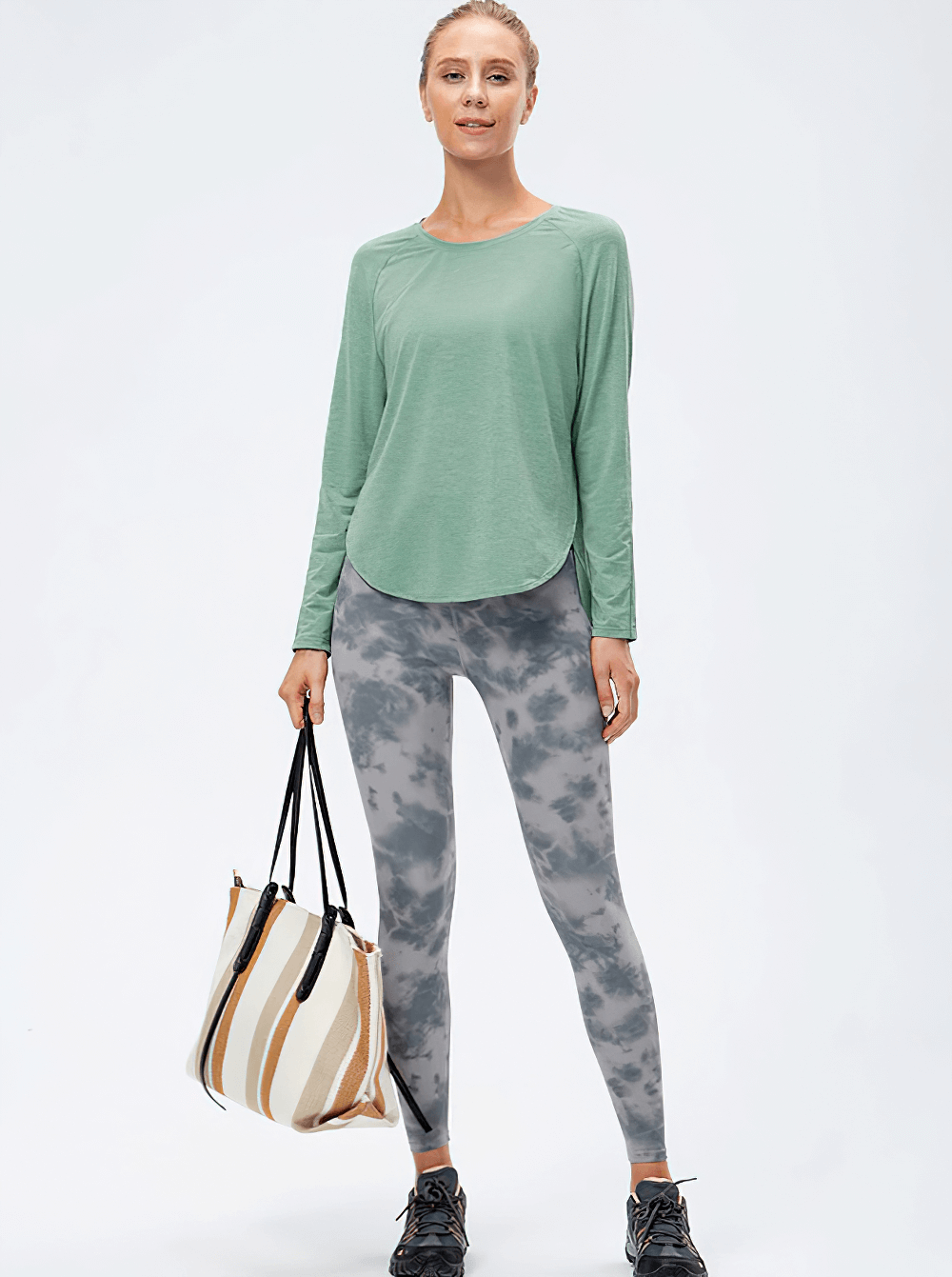 Woman wearing an Elasticity Loose Top with Draped Hem and Split Design, perfect for yoga, paired with gray leggings, holding a striped bag.