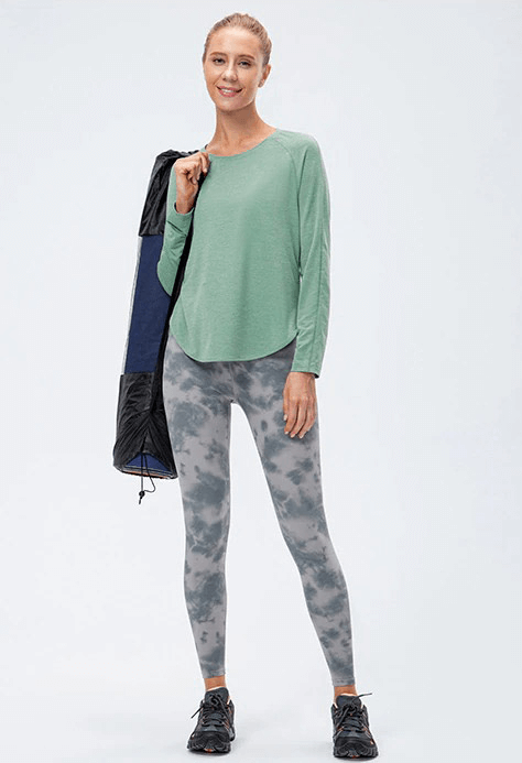 Woman wearing a green long-sleeve draped hem sports top for yoga, paired with patterned leggings, holding a yoga mat.