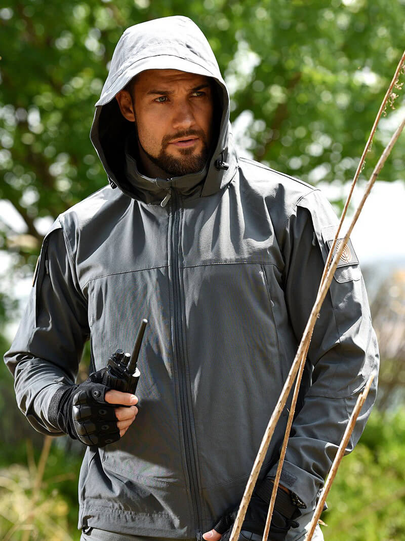 Man wearing grey fashion tactical jacket outdoors, holding a walkie-talkie, perfect for hiking and camping adventures.