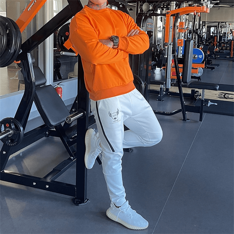 Man in orange sweatshirt and white joggers standing in gym, showcasing fashionable loose fit for activewear lovers.