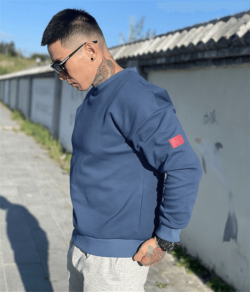 Man wearing fashionable loose blue sweatshirt, sporting casual style with tattoos and sunglasses outdoors.