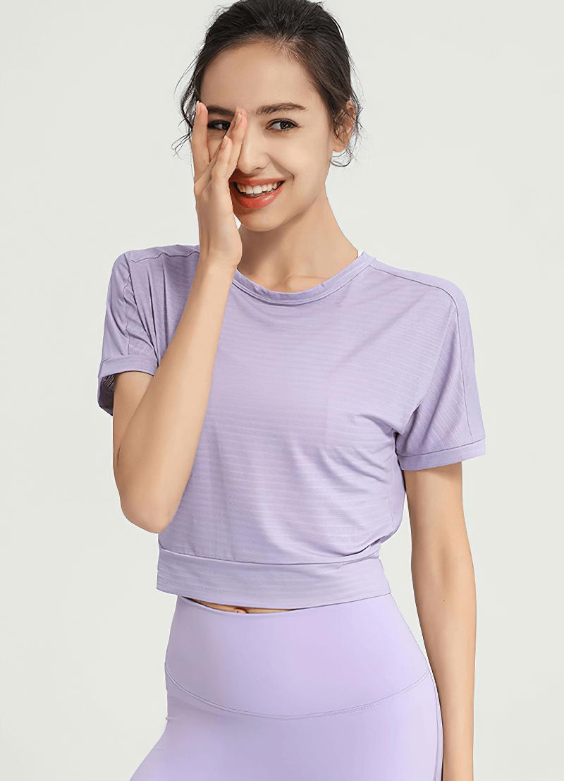 Woman in lavender loose gym crop top, smiling with hand on face, showcasing activewear style.