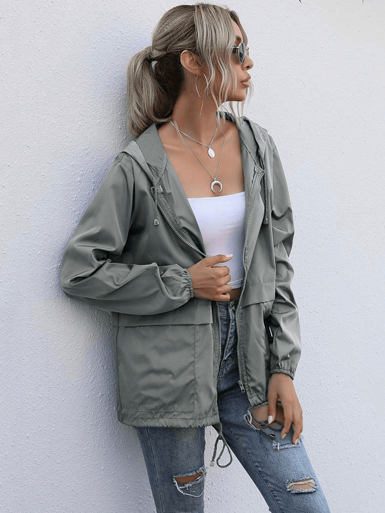 Stylish woman wearing a lightweight waterproof sport jacket with hood, paired with ripped jeans and sunglasses, leaning against a wall.