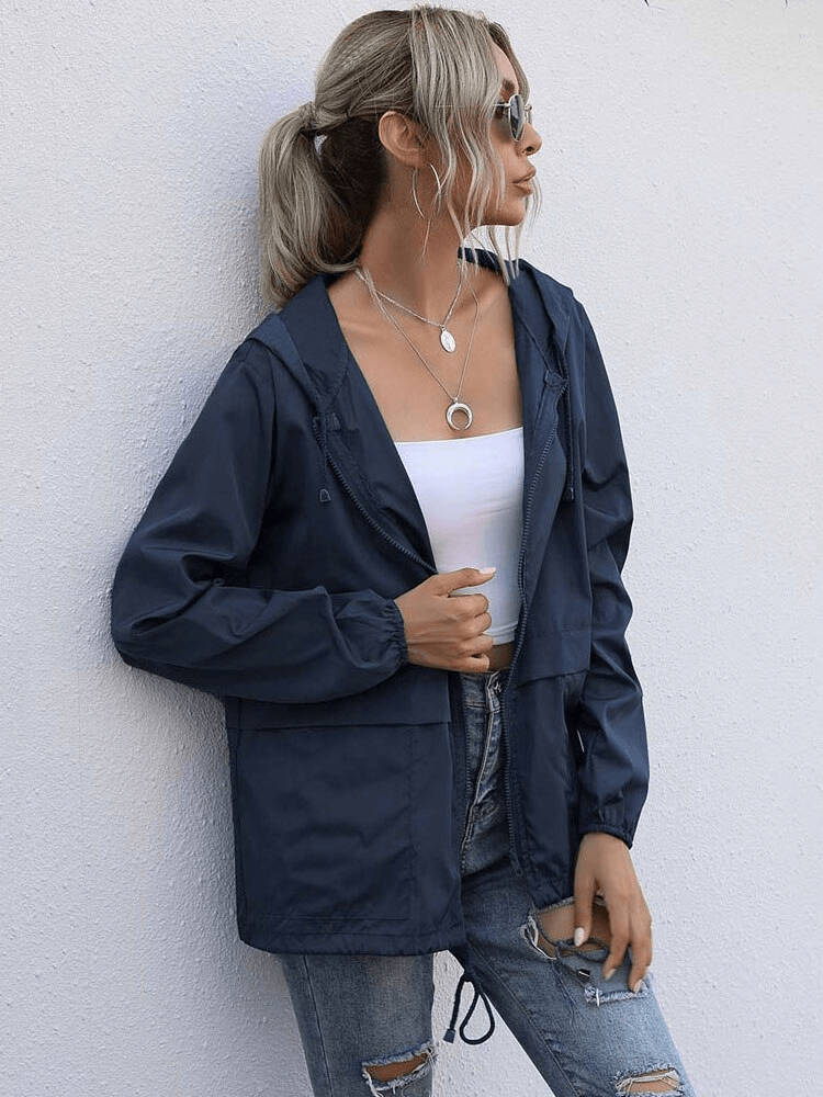 Stylish woman in navy lightweight waterproof sport jacket windbreaker with hood, white top, and jeans, posing against a white wall.