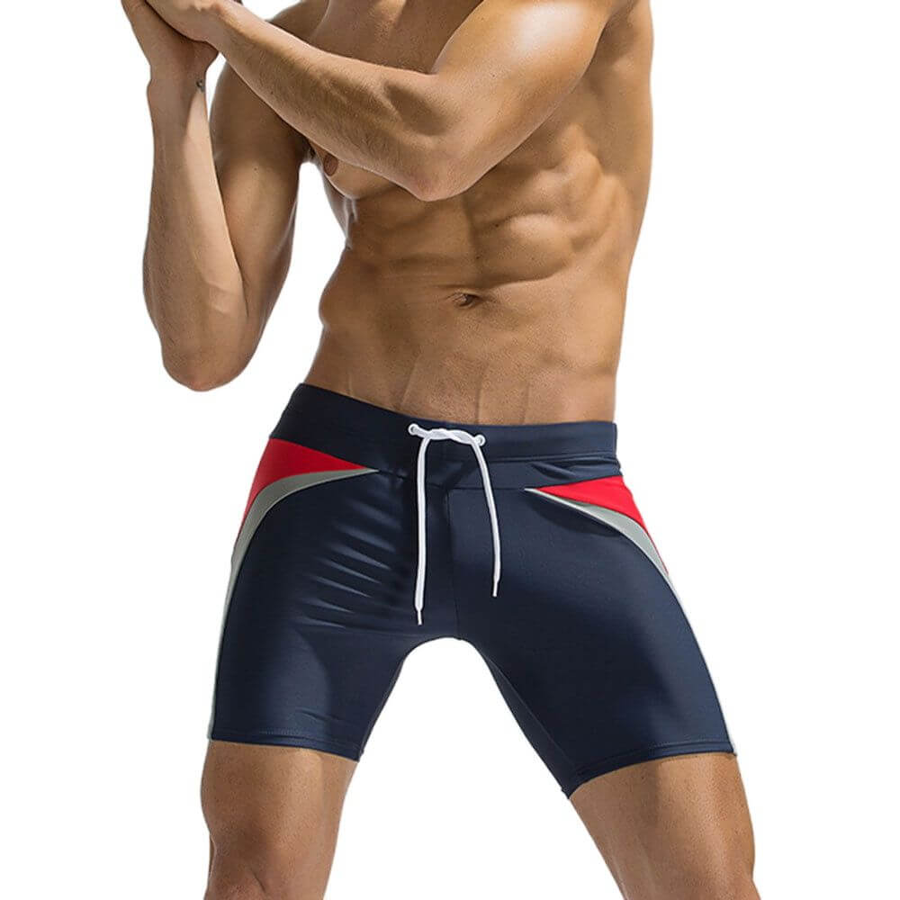 Male model wearing long patchwork swimming trunks in navy with red accents, featuring drawstring closure, perfect for beachwear.