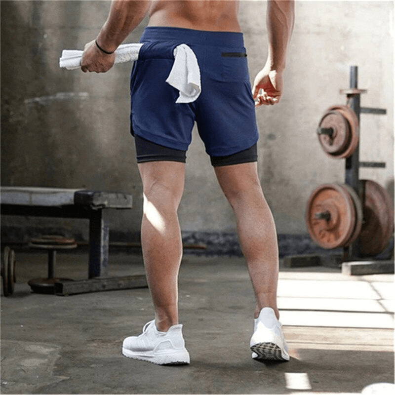 Man wearing blue double-deck running shorts with pockets, ideal for jogging and gym, quick dry, standing in a fitness studio.