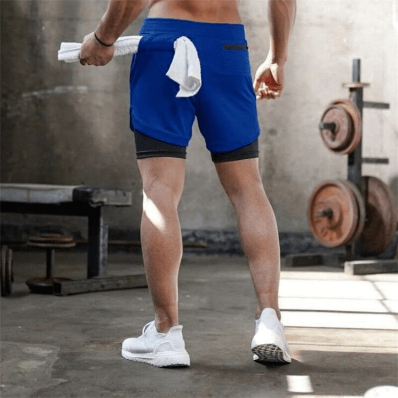 Man wearing blue double-deck running shorts with pockets in gym, showcasing quick-dry fabric, ideal for running and workouts.