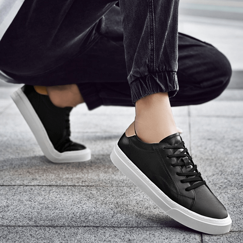 Men's black sporty sneakers with flexible flat sole, lace-up closure, and leather material, worn casually with rolled up pants over a concrete surface.