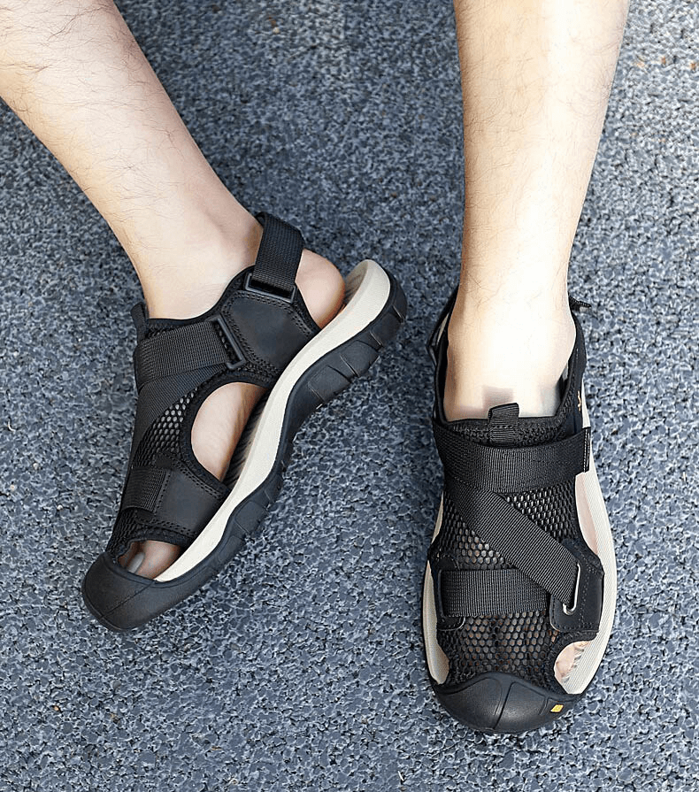 Person wearing black outdoor mesh breathable sandals with Velcro closure on grey surface, showcasing durable design.