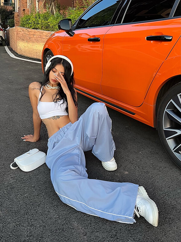 Woman modeling oversized baggy solid high-waisted sweatpants in light blue, sitting by an orange car with white sneakers and a purse.