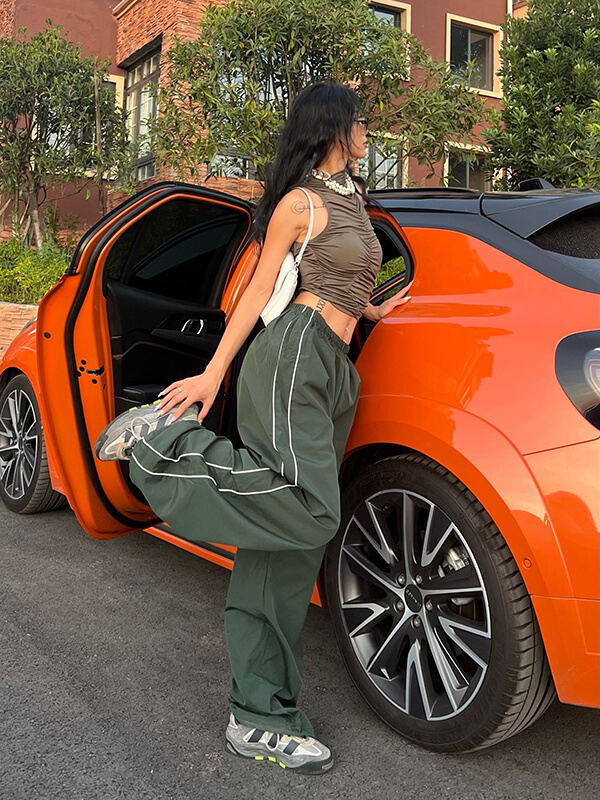 Woman wearing oversized green sweatpants and tiger print top, leaning on orange car, showcasing casual style and comfort.