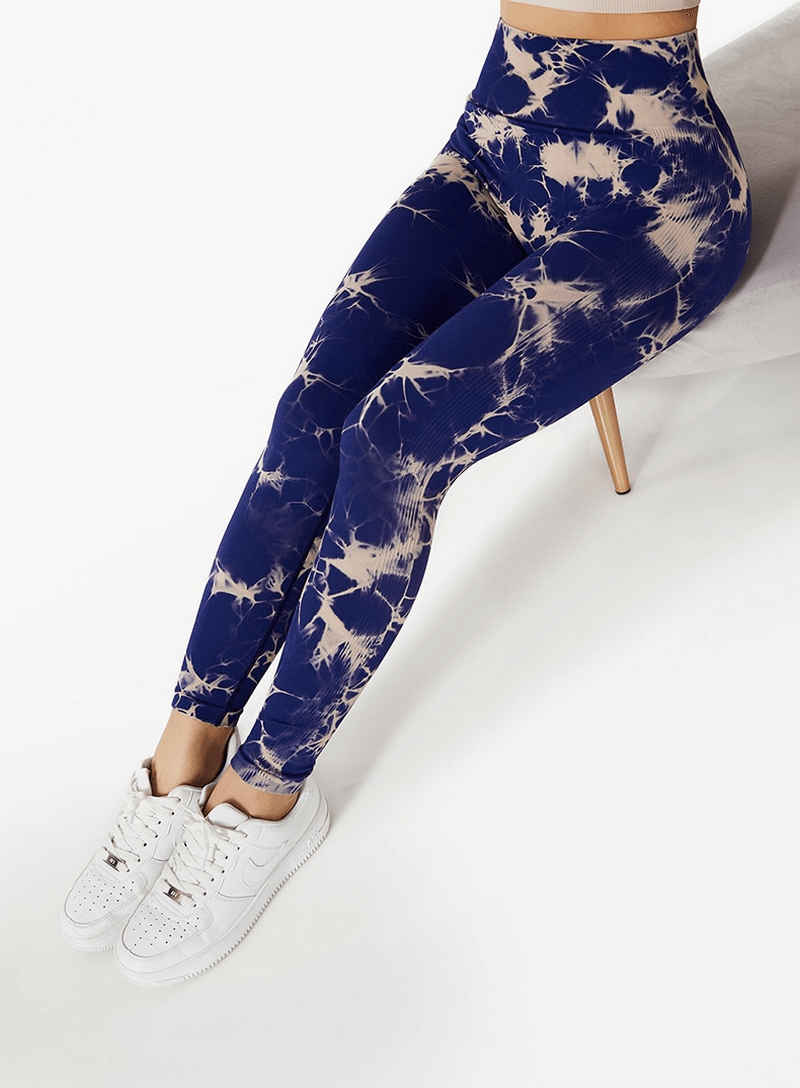 Woman wearing seamless elastic high waist leggings in blue tie-dye pattern, sitting on a chair with white sneakers.