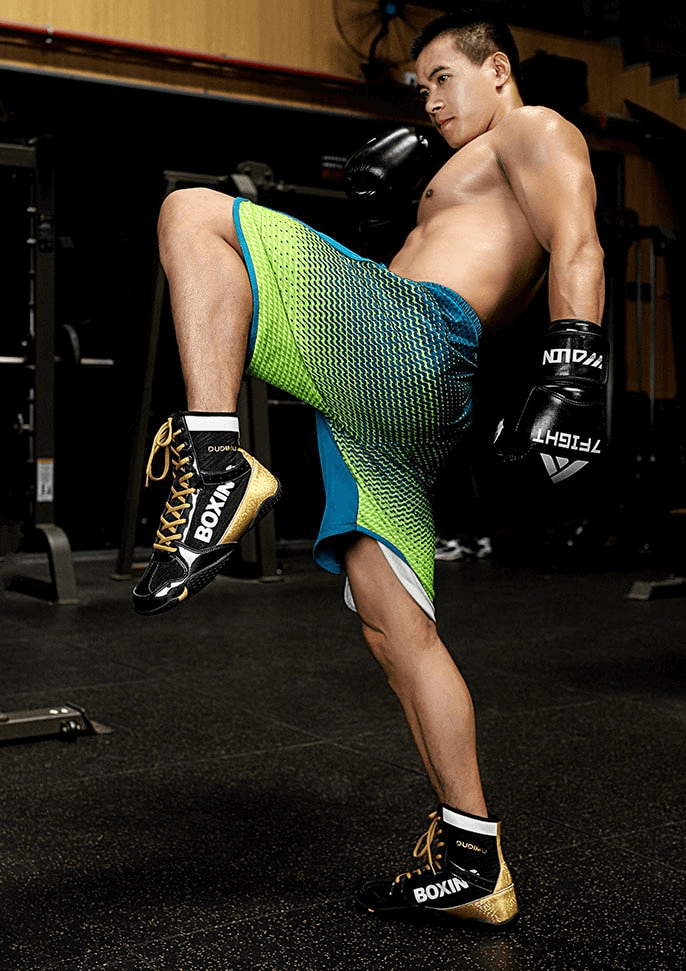 Athlete in boxing gloves and colorful shorts wearing black and gold wrestling sneakers performing a high knee in a gym.