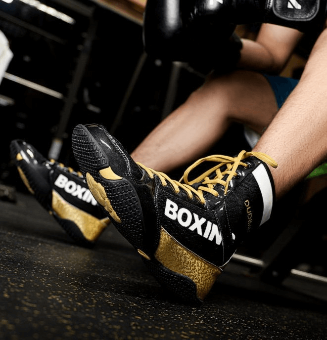 Person wearing black and gold boxing shoes with "BOXING" printed on the side, sitting on a gym floor.