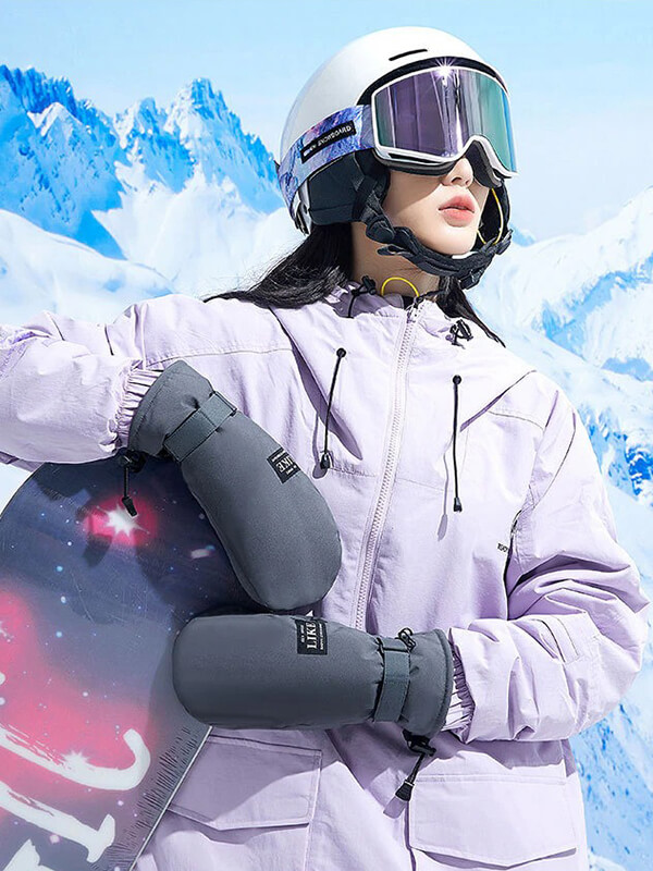 Person wearing skiing gear, holding a snowboard with snowy mountains in the background.
