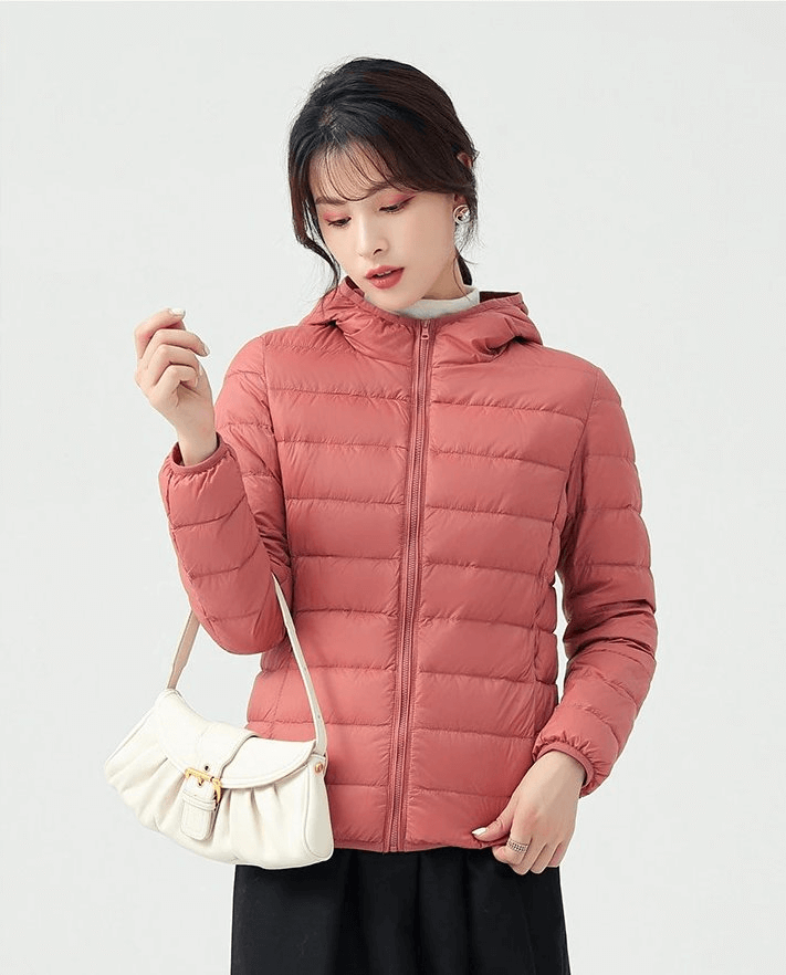 Woman wearing a stylish pink down jacket with a hood, lightweight and insulated, perfect for outdoor activities. Featuring a white handbag.