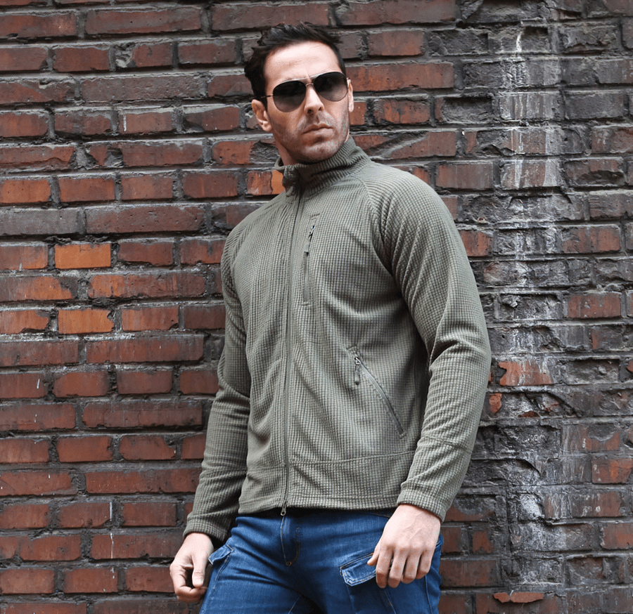 Man wearing tactical lightweight fleece jacket in khaki, standing against a brick wall, showcasing stylish army clothing for men.