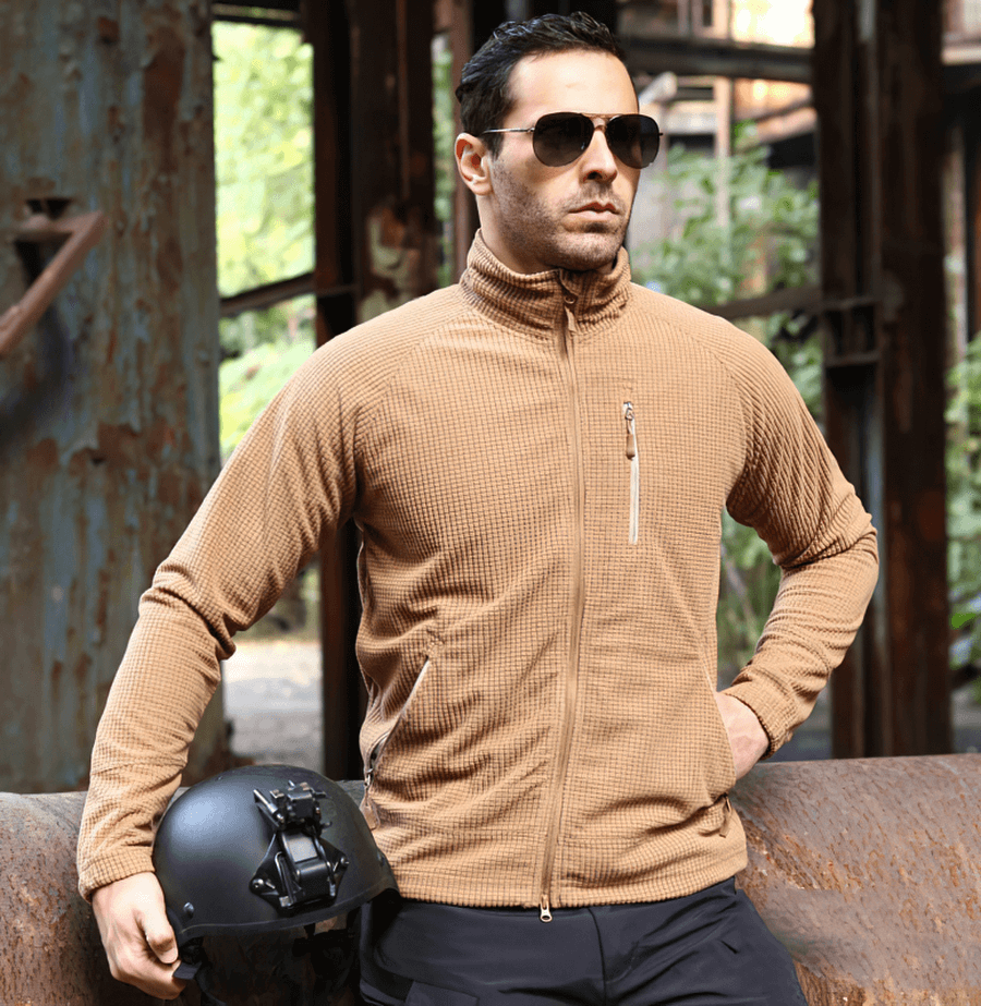 Man wearing Tactical Lightweight Fleece Jacket in khaki, perfect for warmth and style, standing outdoors next to a helmet.
