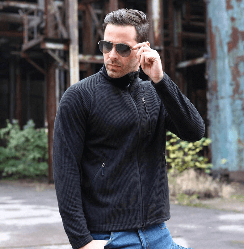 Man wearing black tactical lightweight fleece jacket, standing outdoors in a rugged urban environment.