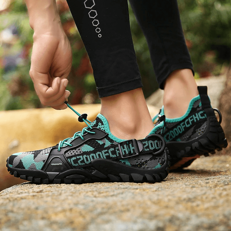 Person wearing Trekking Flexible Hiking Boots with green accents, lacing up on a rocky path. Perfect for outdoor adventures.
