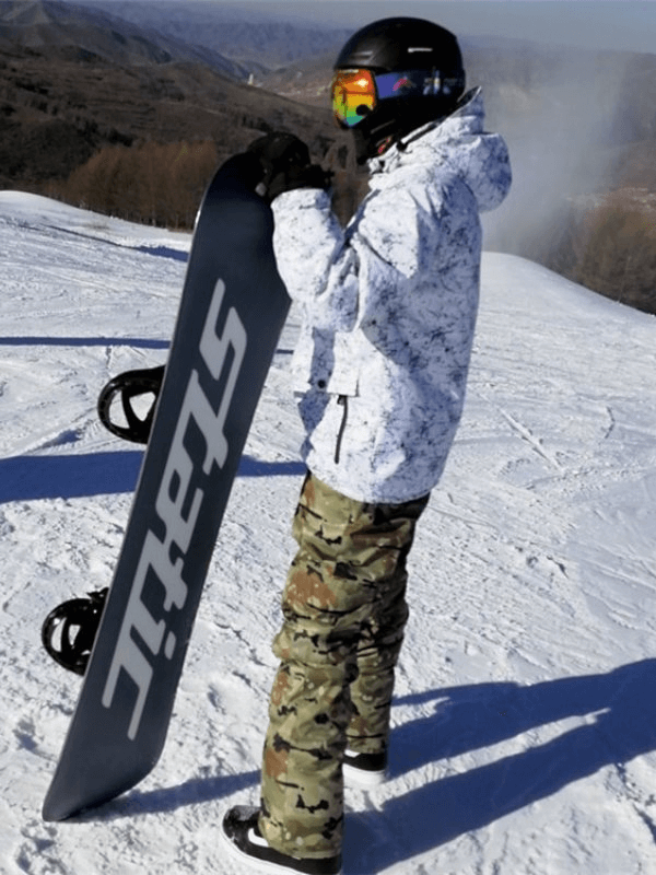 Snowboarder in camo pants and helmet holding a snowboard on a snowy mountain slope.