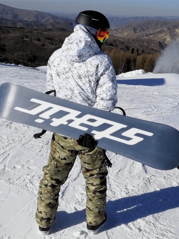 Snowboarder in camouflage pants wearing helmet, holding snowboard on snowy mountain.
