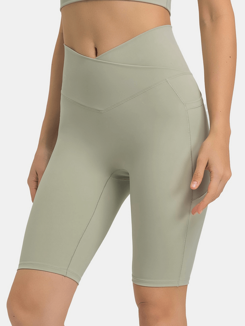 Woman wearing olive green elastic yoga shorts with cross waist and deep pockets, ideal for comfort and support during workouts.