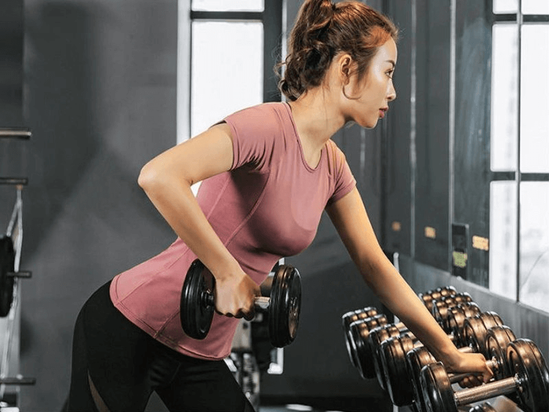 Woman exercising in a quick-dry short sleeve sports T-shirt with transparent back, holding dumbbells in a gym.